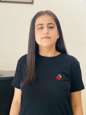 Düz beyaz arka plan önünde duran Berfin Geldi, uzun, düz ve koyu renkli saçlı. Siyah tişört giymiş ve tişörtün sol göğüs kısmında küçük, kırmızı bir kiraz deseni bulunuyor. Doğrudan kameraya bakıyor, yüzünde hafif bir gülümseme var. Arka planda sol üst köşede çerçeveli bir tablo veya fotoğrafın köşesi görünüyor.