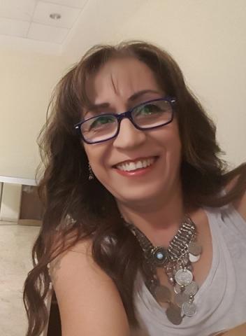 Kahverengi dalgalı saçları, kâkülleriyle, koyu renk çerçeveli gözlüğü ve etnik kolyesi ile içten gülümsüyor.