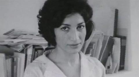 Kısa, siyah, dalgalı saçları, beyaz bluzuyla kitaplarla dolu bir rafın önünde ciddiyetle bakıyor. (Siyah-beyaz fotoğraf)