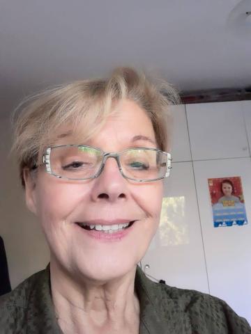 Açık sarı saçlarını toplamış, kâküllerini yana taramış, açık yeşil çerçeveli gözlüğü var. İçten gülümsüyor.