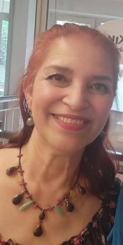 Uzun, kızıl-kahverengi saçlı ve saçları arkaya doğru toplanmış. Yüzünde hafif bir makyaj bulunuyor; gözlerinde eyeliner ve pembe tonlarında bir ruj sürülmüş. Gülümsüyor. Üzerinde çiçek desenli bir bluz ve boynunda renkli taşlardan oluşan gösterişli bir kolye var. Sol kulağında sarkık bir küpe takılı. Arka planda cam bir pencere ve dışarıda ağaçlar görünüyor.