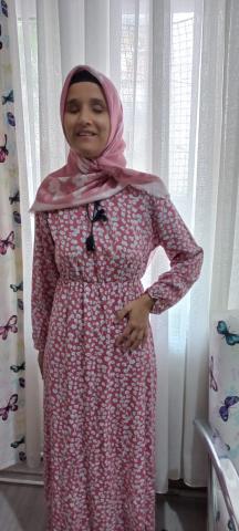 Pembe ve beyaz renkli çiçek desenli bel kısmında siyah bir kemer olan elbise giymiş, başında pembe bir eşarp var. Beyaz tül perde önünde duruyor ve hafif sağa dönük bir şekilde gülümsüyor. Arka planda sol tarafta kelebek desenli bir perde ve sağ tarafta bir pencere görünüyor.