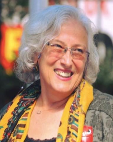 Beyaz, dalgalı, küt saçları; çerçevesiz gözlükleri ile başını soluna çevirmiş, kocaman gülümsüyor. Haki yapraklı bluzunun üzerinde sarı ağırlıklı desenli şalı ve kolyesi var.