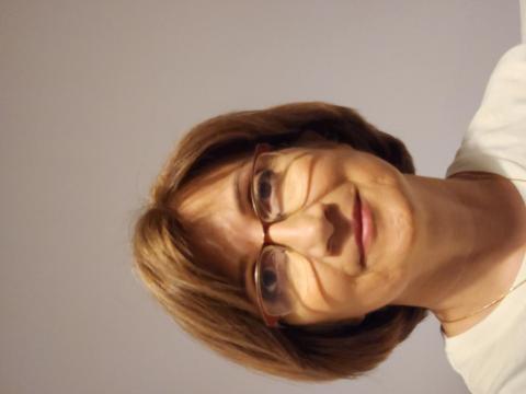 Kısa açık kahverengi saçlı, gözlük takan, açık tenli Nalan Yılmaz duvar önünde duruyor. Beyaz bir üst giymiş ve hafifçe gülümsüyor. Arka plan sade, açık renkli ve herhangi bir detay yok. Yılmaz doğrudan kameraya bakıyor.