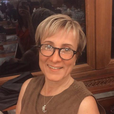 Orta yaşlı, kısa sarı saçlı, siyah gözlüklü, kahverengi bir üst giymiş ve gülümsüyor. Boynunda bir kolye var.