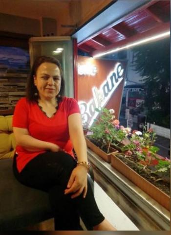 Ve yaka kırmızı tişörtü, siyah pantolonuyla sol eli dizinin üstünde bacak bacak üstüne atmış, bir mekânın çiçekli penceresi önünde gülümseyerek oturuyor.