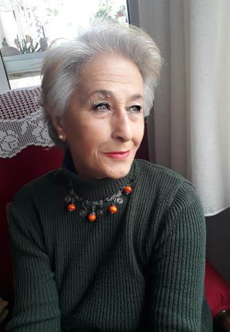 Kısa ve beyaz saçları, kolyesi ve mavi gözleriyle uzaklara bakıyor.