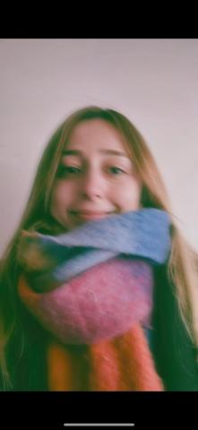 Zeynep Selcen Toprak’ın yüzünde hafif bir gülümseme var. Saçları açık kahverengi ve omuzlarına dökülüyor. Boynuna renkli bir atkı dolanmış, atkıda mavi, gri, pembe ve turuncu gibi canlı renkler var.