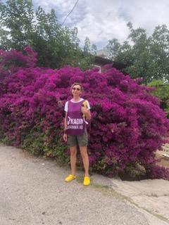 Pembe çiçeklerin önünde ayakta duran bir kadın. Deprem bölgesi Hatay’da bir mahallede, üzerinde Mor Kadın Savunma Önlüğü var.