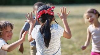 Açık havada çimlerin üzerinde oynayan dört çocuk. Ortadaki çocuk, sırtı kameraya dönük şekilde duruyor ve ellerini havaya kaldırmış. Siyah saçları var ve kırmızı bir bandana takıyor. Diğer üç çocuk ona doğru bakıyor ve gülümsüyorlar. Çocuklar, neşeli bir şekilde oyun oynuyor. Arka planda bulanık bir şekilde ağaçlar ve geniş bir çimenlik alan var.