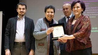 Soldan sağa doğru, genç bir adam, orta yaşlı Gül Erdost , yaşlı bir adam ve başka bir kadın bulunuyor. Genç adam koyu renk bir ceket ve açık renk bir gömlek giymiş. Yanındaki Erdost gri bir hırka giymiş ve elinde bir plaket tutuyor. Yaşlı adam gözlüklü ve takım elbise giymiş. En sağdaki kadın bordo renkli, desenli bir bluz giymiş ve plaketin diğer ucunu tutuyor. Grup, bir ödül töreninde ve arkalarında bir sunum ekranı var. Herkes gülümsüyor ve mutlu.