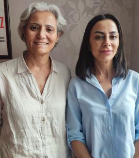 Söyleşideki Sultan Çamur Karataş ve Ceren Ay yan yana duruyor. Soldaki, kısa gri saçlı ve beyaz bir gömlek giymiş. Sağdaki, düz, omuz hizasında siyah saçlı ve açık mavi bir gömlek giymiş. İkisi de kameraya bakarak gülümsüyor. Arka planda, duvarda çiçek desenleri var. Sol üst köşede bir çerçeve içinde yazı bulunan bir posterin bir kısmı görünüyor.