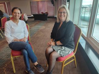 Yazarımız ve konuğu karşılıklı sandalyelerinde oturuyor. Yazarımızın üstünde beyaz bir üst ve kot pantolon var ve gülümsüyor. Konuğumuz sarı saçları ve yüzünde gülümsemesiyle yazarımızın karşısında, üstünde de siyah bluz ve desenli bir etek var. 