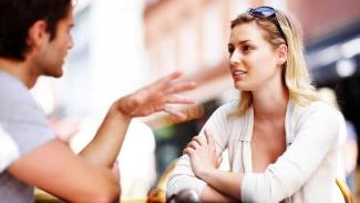 Dış mekânda bir kafede oturan iki kişi. Solda, kısa kahverengi saçlı bir erkek, sağda sarı saçlı bir kadın var. Kadın, kollarını masanın üzerinde çaprazlamış bir şekilde oturuyor ve yüzünde hafif bir gülümseme var. Erkek, ellerini hareket ettirerek konuşuyor. Kadının başının üzerinde güneş gözlüğü var.