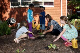 Açık havada, bahçede 8-10 yaşlarında birkaç çocuk ve bir yetişkinle birlikte çiçek ekiyorlar.