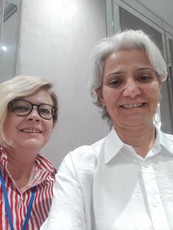 Söyleşideki iki kadın. Soldaki sarı saçlı ve gözlük takıyor. Üzerinde kırmızı ve beyaz çizgili bir gömlek var. Sağdaki kısa, beyaz saçlı ve beyaz bir gömlek giymiş. İkisi de kameraya bakarak gülümsüyor. Arka planda büyük bir ayna ve açık renkli bir duvar görülüyor.