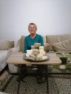 Salonda büyük koltukta düz beyaz kısa saçlı, gözlüklü orta yaşlı kadın oturuyor. Kadının önünde bir masa var. Masada büyük bir servis tabağında beyaz ve krem renginde birçok kalıp sabun var. Kadın gülümseyerek karşı tarafa bakıyor.