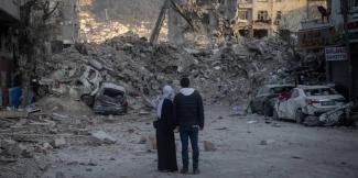 Yıkılmış binaların ve enkazın bulunduğu bir sokak. İki kişi, bir kadın ve bir erkek, sırtları kameraya dönük şekilde enkaza bakıyorlar. Kadın siyah uzun bir elbise ve beyaz bir başörtüsü, erkek siyah bir mont ve kot pantolon giymiş. Sokakta, enkazın arasında sıkışmış ve hasar görmüş araçlar var. Arka planda dağlık bir alan var.