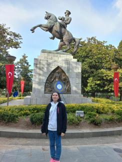 Fatma Aykın, Atatürk'ün at üstünde olduğu bir heykelin önünde duruyor. Beyaz bir kapüşonlu sweat tişört ve siyah bir ceket giymiş, mavi kot pantolon ve pembe spor ayakkabılarla tamamlamış. Heykelin kaidesi taş bloklardan yapılmış ve üzerinde bir çelenk yer alıyor. Arka planda yeşil ağaçlar ve Türk bayrakları görülüyor. Gökyüzü açık ve güneşli.