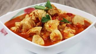 Beyaz bir kâsede servis edilen tavuk yemeği. Tavuk parçaları, kırmızı biber ve domates sosu ile pişirilmiş. Üzerinde birkaç dal maydanoz yaprağı ile süslenmiş. Arka planda ahşap bir masa yüzeyi görülüyor. Kâse, beyaz bir tabak üzerinde duruyor.