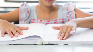 Bir çocuk, Braille alfabesiyle yazılmış bir kitabı okuyor. Çocuğun elleri kitabın üzerinde ve parmaklarıyla sayfaları hissediyor. Üzerinde kısa kollu, desenli bir elbise var.