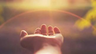 Açık bir avuç içi yukarı doğru bakıyor. Arka planda, gün batımı veya doğuşu gibi görünen sıcak tonlarda bir ışık parlıyor. Işık, avuç içine doğru vuruyor ve etrafında hafif bir hale oluşturuyor. Arka plan bulanık ve doğa manzarası gibi görünüyor.