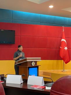 Bir konferans salonunda, kürsüde duran Özge Sarıkaya, koyu gri bir kıyafet giymiş ve elinde mikrofon tutuyor. Kürsünün önünde bir logo var. Arka planda kırmızı ve mavi renkli panellerle kaplı bir duvar bulunuyor. Duvarın sağ tarafında bir Türk bayrağı asılı. Kürsünün önünde, masanın üzerinde beyaz kağıtlar ve kırmızı kapaklı kitaplar yer alıyor. Tavanda yuvarlak bir ışık yanıyor. Sol üst köşede kapalı bir televizyon ekranı var.