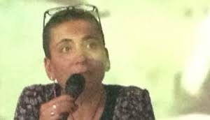 Kısa saçlı Hülya Onur Yaman, elinde mikrofon tutuyor. Üzerinde koyu renkli bir kıyafet var ve gözlükleri başının üstünde duruyor. Arka plan bulanık ve açık renkli. Yaman, bir şeyler anlatıyor.