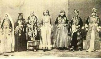 Yedi kadın, geleneksel kıyafetler içinde yan yana duruyor. Her biri farklı desen ve süslemelerde uzun elbiseler giymiş. Başlarında çeşitli örtüler ve aksesuarlar bulunuyor. Kıyafetler genellikle uzun kollu ve bol kesimli. Kadınlar, farklı yöresel kıyafetleri temsil ediyor. Arka planda sade bir perde var ve kadınlar halı üzerinde duruyor.