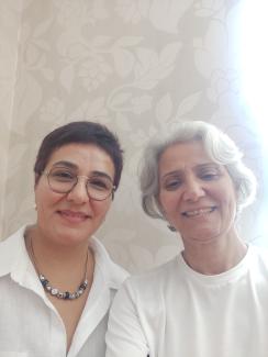 İki kadın yan yana duruyor ve kameraya gülümsüyor. Soldaki kısa koyu renk saçlı, gözlük takmış ve beyaz bir gömlek giymiş. Boynunda boncuklu bir kolye var. Sağdaki kısa gri saçlı ve beyaz bir tişört giymiş. Arka planda açık renkli, desenli bir duvar kâğıdı var. Işık sağ taraftan geliyor ve fotoğrafın sağ tarafında parlak bir alan oluşturmuş.