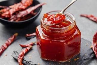 Altıgen bir cam kavanozda kırmızı biber reçeli veya sosu bulunuyor. Kavanozun içinden bir kaşıkla biraz sos alınmış. Kavanozun etrafında kurutulmuş kırmızı biberler var. Arka planda siyah bir kâsede daha fazla kurutulmuş kırmızı biberler görülüyor.