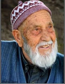 Yaşlı bir adam gülümseyerek sağ tarafa bakıyor. Beyaz sakalı ve bıyığı var. Başında kırmızı ve beyaz desenli bir takke bulunuyor. Üzerinde mavi bir ceket ve koyu renkli bir gömlek giymiş. Yüzünde yaşlılığa bağlı kırışıklıklar ve lekeler mevcut. Arka plan bulanık, bu da adamın yüzüne odaklanmayı sağlıyor.