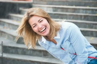 Merdivenlerde duran genç bir kadın, başını yana eğmiş ve içten bir şekilde gülüyor. Saçları açık ve omuz hizasında. Üzerinde mavi beyaz çizgili bir gömlek var. Arka planda bulanık merdiven basamakları görülüyor. Yüz ifadesi oldukça neşeli ve samimi.