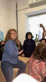 Bir odada bir grup insan var. Önde, mavi gömlek ve kot pantolon giymiş, gözlüklü, kıvırcık saçlı bir kadın, ahşap bir masanın kenarına oturmuş ve gülümsüyor. Arkasında, koyu renkli giysiler giymiş birkaç kadın daha var. Duvarın önünde, pencere kenarında birkaç seramik vazo ve dekoratif bir at figürü bulunuyor. Arka planda, duvarda bir afişin bir kısmı görünüyor. Kadınlar, samimi bir ortamda sohbet ediyor.