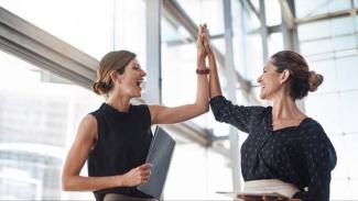 İki kadın, modern bir ofis ortamında birbirlerine "çak" yaparak sevinçlerini paylaşıyor. Biri siyah kolsuz bir bluz giymiş ve elinde bir dosya tutuyor. Diğeri koyu renkli, desenli bir bluz giymiş ve elinde bir tablet veya defter tutuyor. İkisi de gülümsüyor ve oldukça mutlu görünüyorlar. Arka planda büyük cam pencereler ve modern bir bina yapısı dikkat çekiyor.