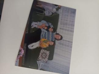 Yeşil bir koltukta oturan üç kişi. Ortada bir kadın, kucağında küçük bir çocuk var. Kadın mavi-beyaz çizgili bir kazak giymiş. Küçük çocuk beyaz çorap ve mavi bir kıyafet giymiş. Kadının solunda, sarı bir elbise giymiş başka bir çocuk oturuyor. Arka planda beyaz tül perdeler ve bir sehpa üzerinde çiçekler var. Fotoğrafın sol alt köşesinde de çiçekler görülüyor.