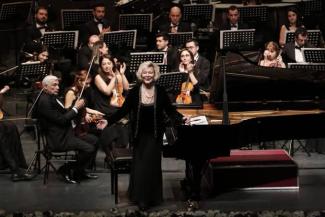 Bir konser salonu. Ön planda, siyah elbise giymiş Gülsin Onay, piyanonun yanında duruyor ve izleyicilere doğru gülümsüyor. Arkasında, çeşitli enstrümanlar çalan bir orkestra. Orkestra üyeleri siyah takım elbise giymiş. Bazı müzisyenler keman çalarken, diğerleri farklı enstrümanlarla meşgul. Arka planda, notaların yer aldığı notalıklar bulunuyor. Orkestra üyelerinin çoğu Onay’ı alkışlıyor. Sahnenin ön kısmında çiçeklerle süslenmiş bir alan bulunuyor.