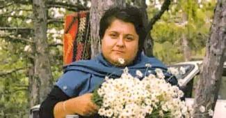 Ağaçların arasında oturan Kutsiye Bozoklar, elinde büyük bir papatya demeti tutuyor. Üzerinde mavi bir şal var, gülümsüyor ve arka planda bir araba ve yeşil yapraklı ağaçlar dikkat çekiyor.