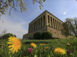 Anıtkabir, çimenlerin ve çiçeklerin arasında yükselen görkemli bir yapı olarak görülüyor. Ön planda sarı ve pembe çiçekler, yapının etrafında ağaçlar ve yeşillikler bulunuyor. Gökyüzü açık ve mavi, birkaç beyaz bulut var. Büyük sütunlar ve taş duvarlar dikkat çekiyor.