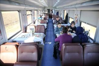 Tren vagonunda yemekli bir bölüm. Masalarda beyaz örtüler. Bazı masalarda insanlar oturuyor. Ön planda, üç kadın ve bir çocuk masada yemek yiyor. Kadınlardan biri mor bir ceket, diğerleri koyu renkli giysiler giymiş. Arka planda başka masalarda da oturan insanlar var. Vagonun pencerelerinden dışarısı görünüyor, tren hareket halinde.