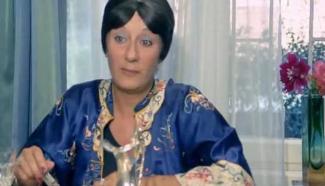 Sevda Aydan mavi ve desenli bir sabahlık giymiş, masada oturuyor. Kısa siyah saçları var ve yüzü ciddi bir ifade taşıyor. Masanın üzerinde bir bardak ve başka cam eşyalar bulunuyor. Arka planda, pencere önünde vazoda çiçekler var. Pencerenin dışında bulanık bir manzara görünüyor.