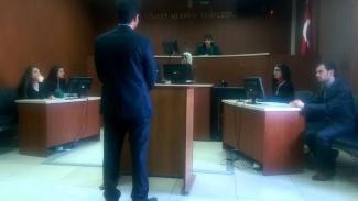 Bir mahkeme salonu. Ortada bir avukat veya savcı ayakta duruyor ve kürsüye doğru bakıyor. Kürsünün arkasında bir hâkim, sağ tarafta iki kişi oturuyor, biri bilgisayar ekranına bakıyor. Sol tarafta iki kişi daha oturuyor, onların da önünde bir bilgisayar var. Duvarın arkasında "Adalet mülkün temelidir." yazısı ve bir Türk bayrağı görülüyor.