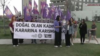 Bir grup insan, bir araya gelmiş ve pankartlar taşıyor. Pankartın üzerinde "DOĞAL OLAN KADINLARIN KARARINA KARIŞMAMAK" yazıyor. Pankartın altında "CİNAYETLERİ DURDURACAĞIZ PLATFORMU" ve "GENÇ FEMİNİSTLER" logoları bulunuyor. Arkada, mor renkli bayraklar taşıyan insanlar var, bayrakların üzerinde kadın sembolleri ve "DOĞAL OLAN NORMA DEMOKRASİ" yazıları yer alıyor. Katılımcılar arasında mikrofon ve megafon kullanan kişiler de var. Arka planda yüksek binalar ve "ANKARA İL SAĞLIK MÜDÜRLÜĞÜ" yazısı var. 