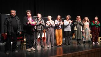 Sahnede bir grup insan yan yana duruyor. Sol başta siyah mont giymiş bir adam, yanında, elinde çiçek buketi tutan ve siyah takım elbise giymiş bir kişi, ortada, deri ceket giymiş yaşlı bir adam, onun yanında, desenli elbise giymiş birkaç kadın sıralanmış. Kadınlardan bazıları ellerini çırpıyor. En sağda, kırmızı yelek ve yeşil bluz giymiş bir kadın var. Sahne arka planı siyah ve sahnede birkaç sandalye bulunuyor.