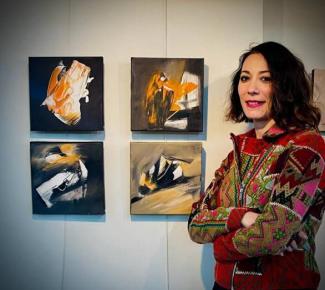 Bir sanat galerisi veya sergi alanı. Kısa saçlı Özge Gökbulut Özdemir, kollarını kavuşturmuş, üzerinde renkli desenli bir ceket var. Arka planda duvarda asılı dört adet küçük kare tablo bulunuyor. Bu tablolar soyut tarzda ve ağırlıklı olarak siyah, beyaz, turuncu ve gri tonlarında. Tabloların önünde durarak kameraya gülümsüyor. 