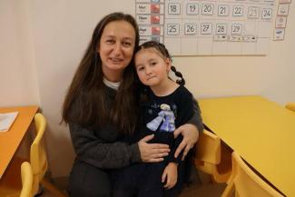 Bir kadın ve küçük bir kız çocuğu sarı masaların bulunduğu bir sınıfta oturuyor. Kadın, uzun kahverengi saçlı ve gri bir kazak giymiş. Kız çocuğu Elsa karakterinin resminin olduğu koyu renkli bir elbise giymiş ve saçları iki yandan örgülü. Arka planda, duvarda bir takvim var ve üzerinde günler ve aylar yazılı. Masalar ve sandalyeler sarı renkte. Kadın, kızı kucağına almış ve ikisi de kameraya bakarak gülümsüyor.