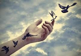 Gökyüzüne uzanan bir el ve elin üzerinde uçan kuşlar, elin bilek kısmında kuş dövmeleri bulunuyor. Gökyüzü bulutlu ve kuşlar özgürce uçuyor.