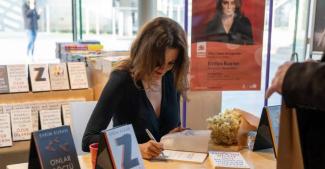 Kitap imza etkinliğinde bir kadın, masada oturmuş ve bir kitabı imzalıyor. Kadının önünde birkaç kitap yığılmış. Masanın üzerinde "Evrim Kuran" ismi yazılı kitaplar ve bir buket çiçek var. Arka planda, kadının büyük bir posteri asılı. Posterde "Evrim Kuran" ismi ve "Onlar Göçtü Buradan" kitabının tanıtımı yer alıyor. Çevrede başka kitaplar da sergilenmiş.