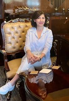 Ayşe Gülçin İlhan, klasik tarzda dekore edilmiş bir odada, şık bir koltukta oturuyor. Önünde, cilalı ahşap bir masa, masanın üzerinde birkaç bardak altlığı ve bir peçete var. Beyaz bir gömlek ve beyaz pantolon giymiş, ayaklarında beyaz spor ayakkabılar var. Saçları kısa ve koyu renkli, gülümsüyor ve ellerini kucağında birleştirmiş. Koltuk, altın renginde ve deri kaplı. Arka planda, ahşap bir dolap ve üzerinde dekoratif objeler yer alıyor. 