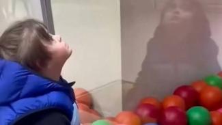 Mavi yelek giymiş bir çocuk, bir camın arkasındaki renkli toplara bakıyor. Camda çocuğun yansıması da görülüyor. Toplar kırmızı, turuncu ve yeşil. Çocuk yukarı doğru bakıyor ve ifadesi meraklı.